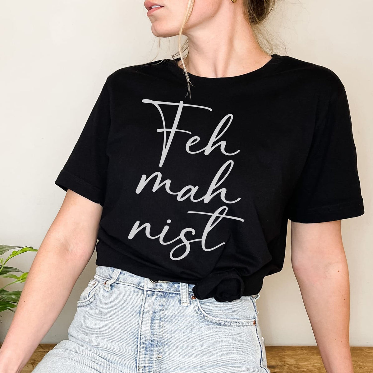 Person wearing a black feminist t-shirt with 'Feh mah nist' text against a neutral background