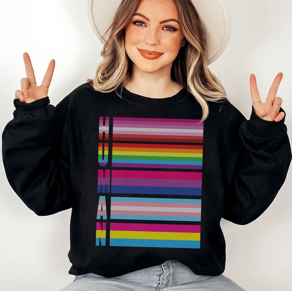 Person wearing a black LGBTQ sweatshirt with a colorful 'HUMAN' pride flags design, making peace signs.