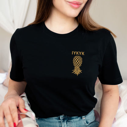Person wearing a black t-shirt with a gold pineapple logo and text: Subtle Swingers Shirt, Upside Down Pineapple Pocket Design Tee, IYKYK T-Shirt, Discreet  Hotwife Tshirt, Gift For Non Monogamy Couples