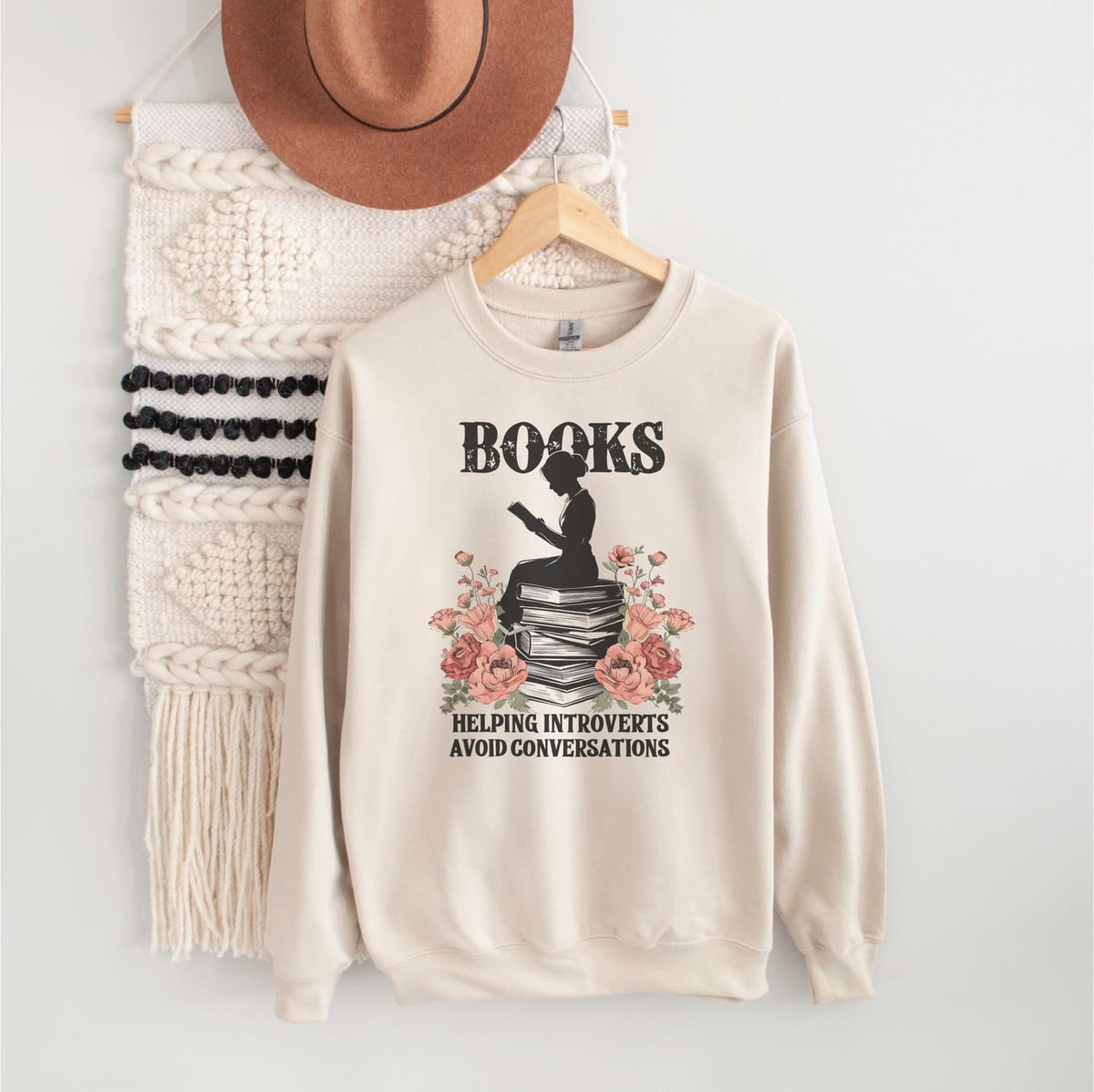 Beige bookish sweatshirt with a graphic design on a hanger against a white wall.