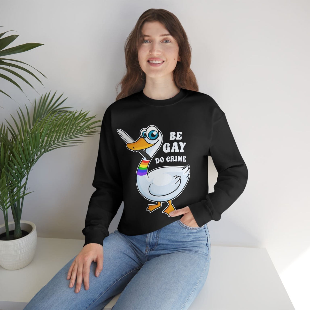 Person wearing a black LGBTQ activist sweatshirt with a graphic design and text, sitting on a white couch with a plant in the background.