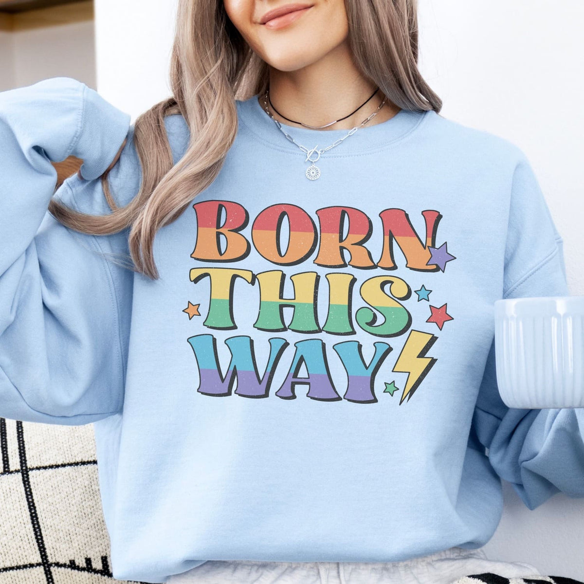 Person wearing a light blue lgbtq pride sweatshirt with 'Born This Way' text in colorful letters.