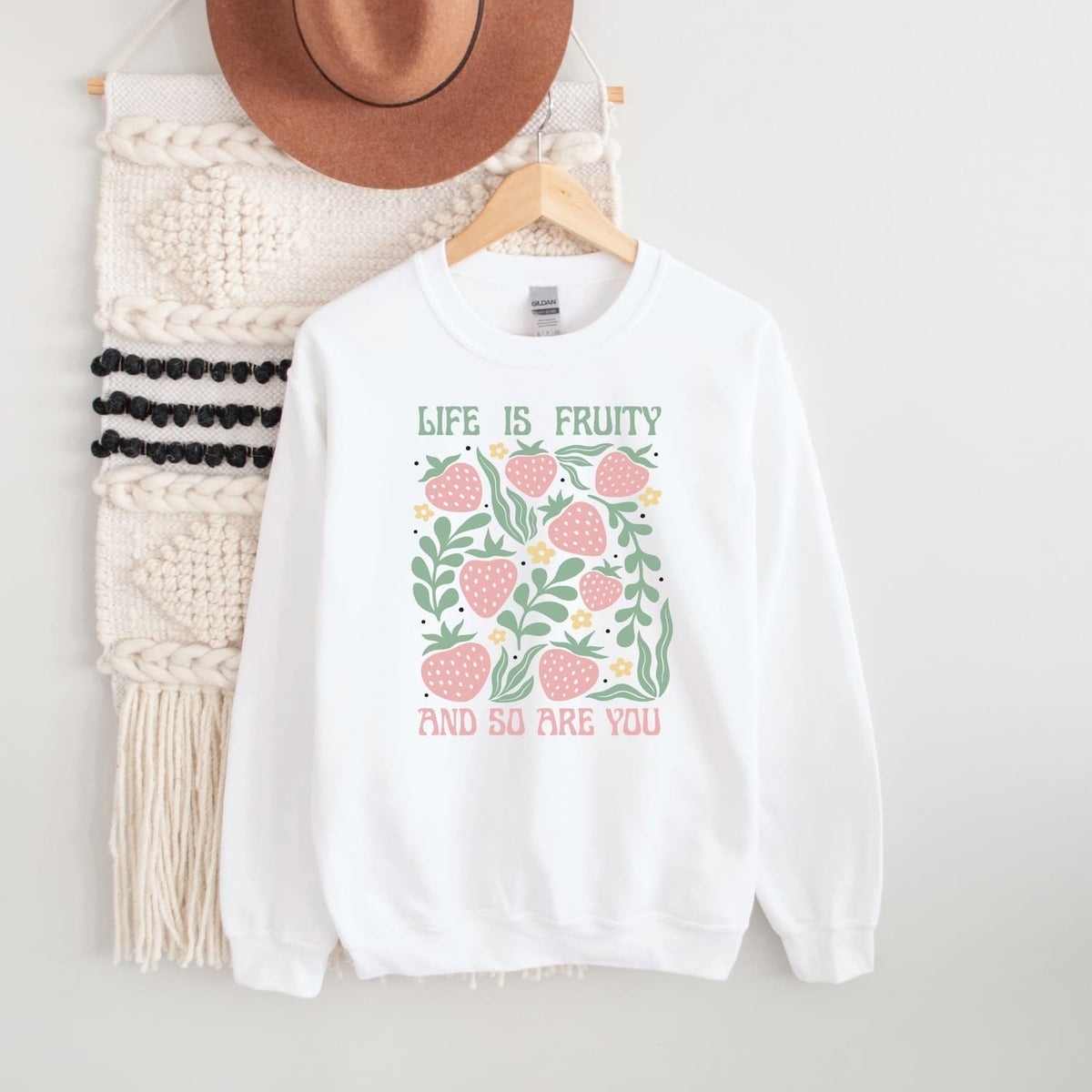 White fruity queer sweatshirt with strawberry design and text on a hanger with a hat and scarf in the background.