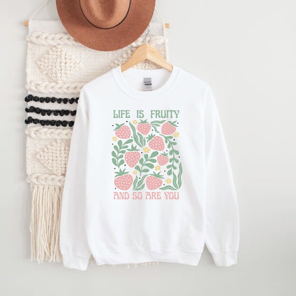 White fruity queer sweatshirt with strawberry design and text on a hanger with a hat and scarf in the background.