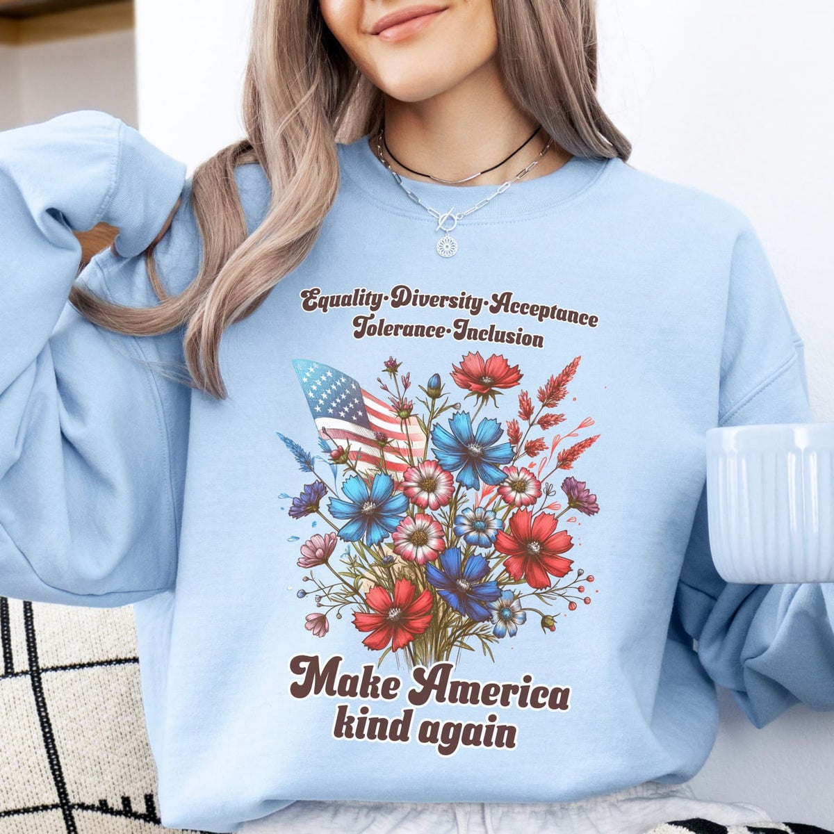 Person wearing a light blue american patriotic sweatshirt with a floral design and text, holding a white mug.