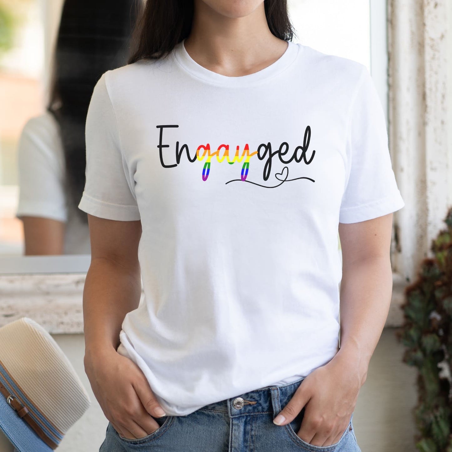 Queer person wearing a white wedding announcement t-shirt with 'Engayged' text in rainbow colors.