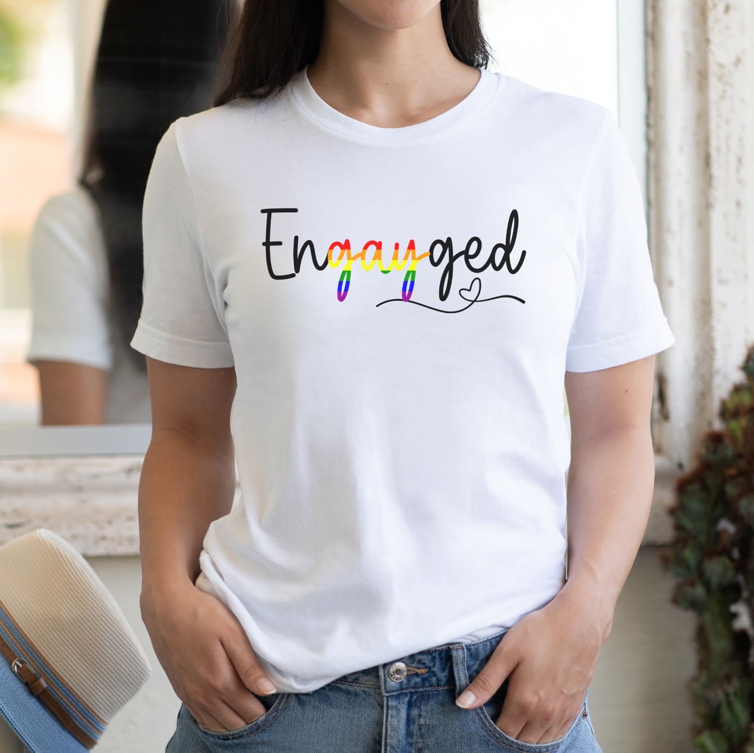 Queer person wearing a white wedding announcement t-shirt with 'Engayged' text in rainbow colors.
