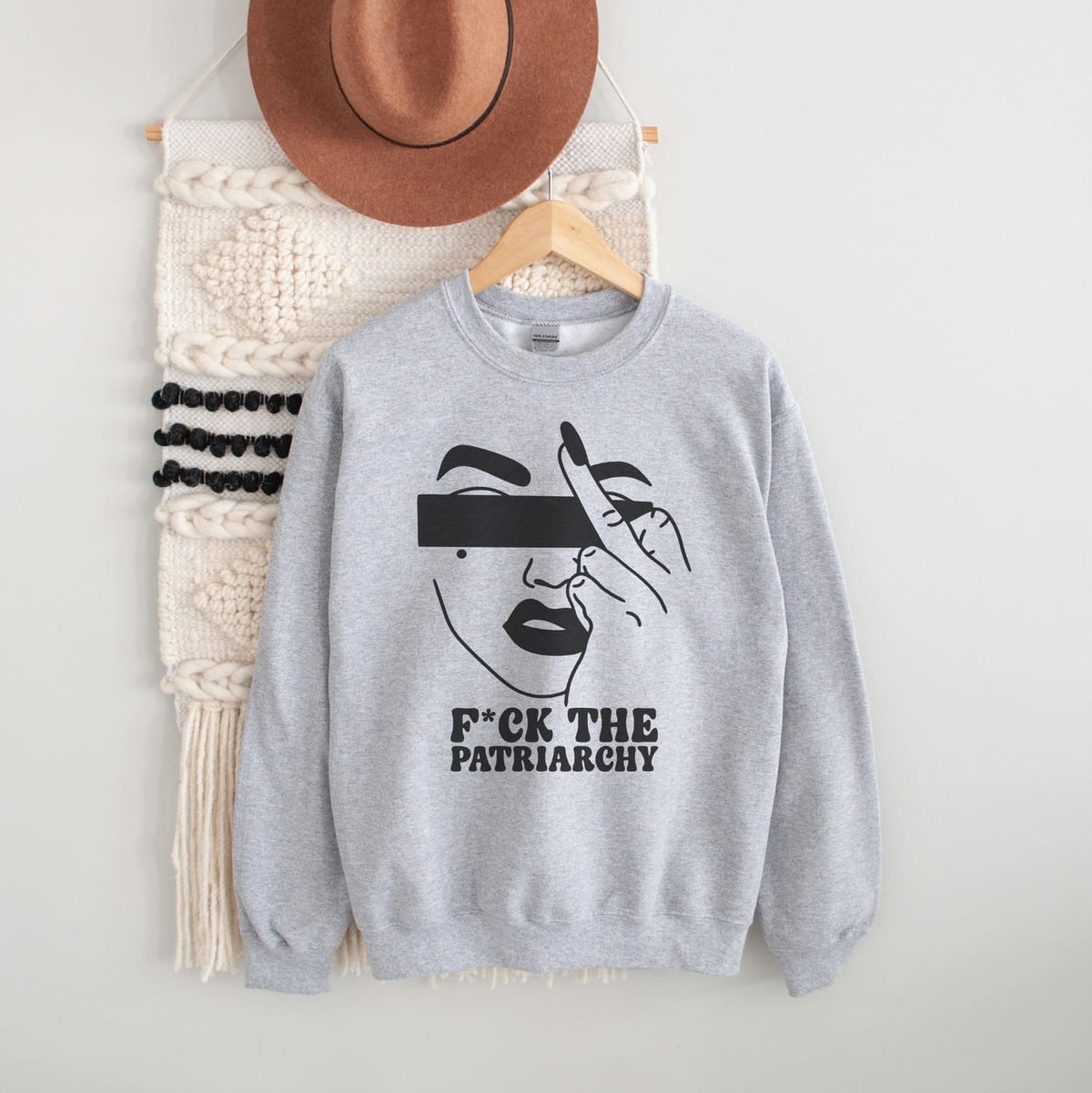 Gray sweatshirt with a feminist graphic design and text, placed on a white surface with a hat and bag in the background.