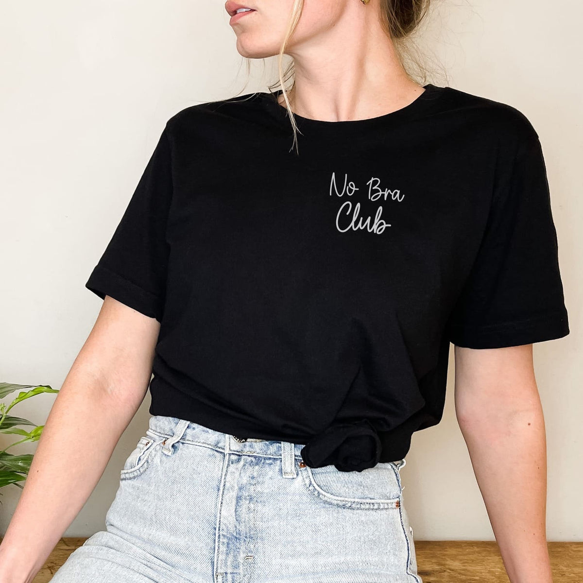 Person wearing a black feminist body positivity t-shirt with 'No Bra Club' text, standing against a neutral background.