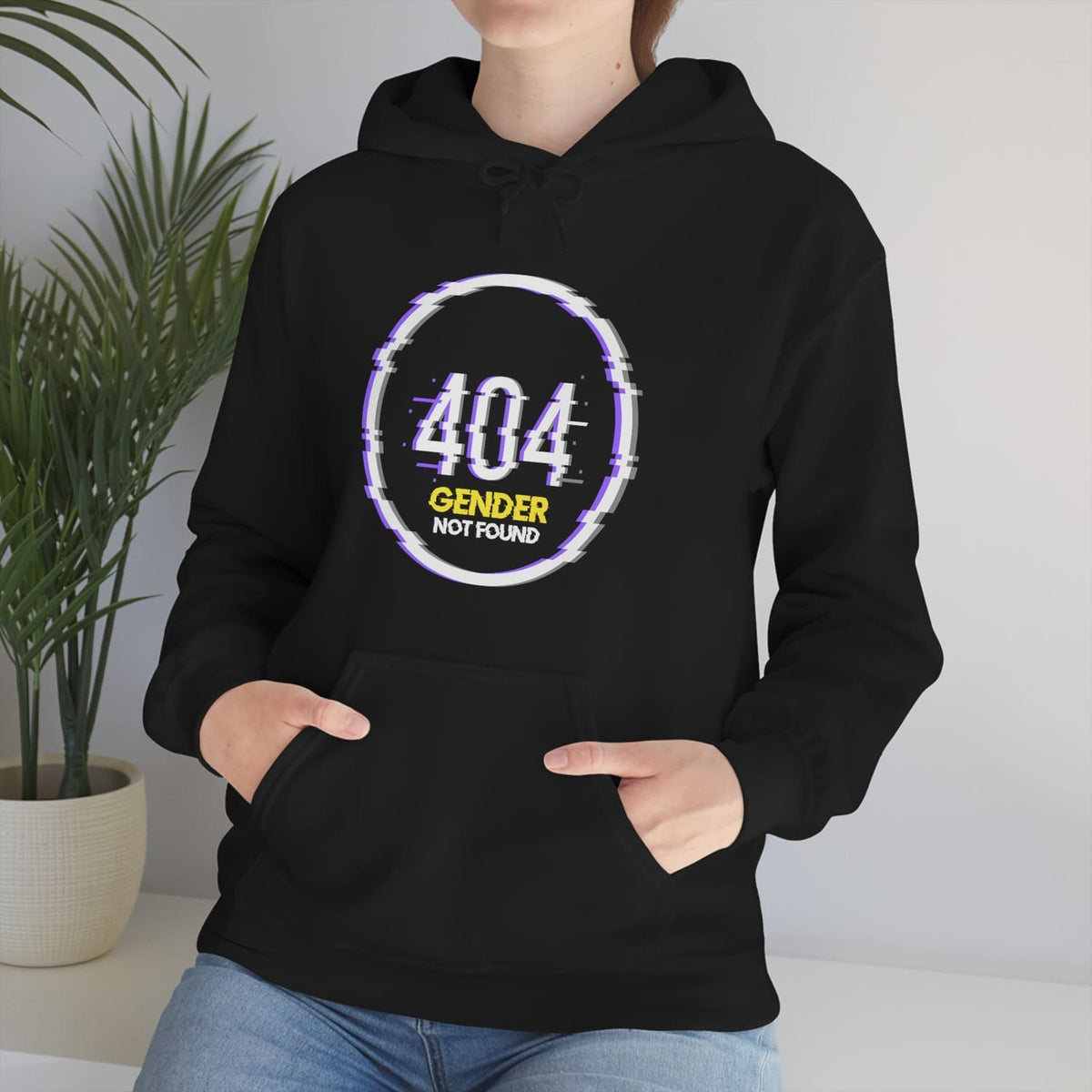Person wearing a black funny nonbinary hoodie with '404 Gender Not Found' design, standing indoors with a plant in the background.