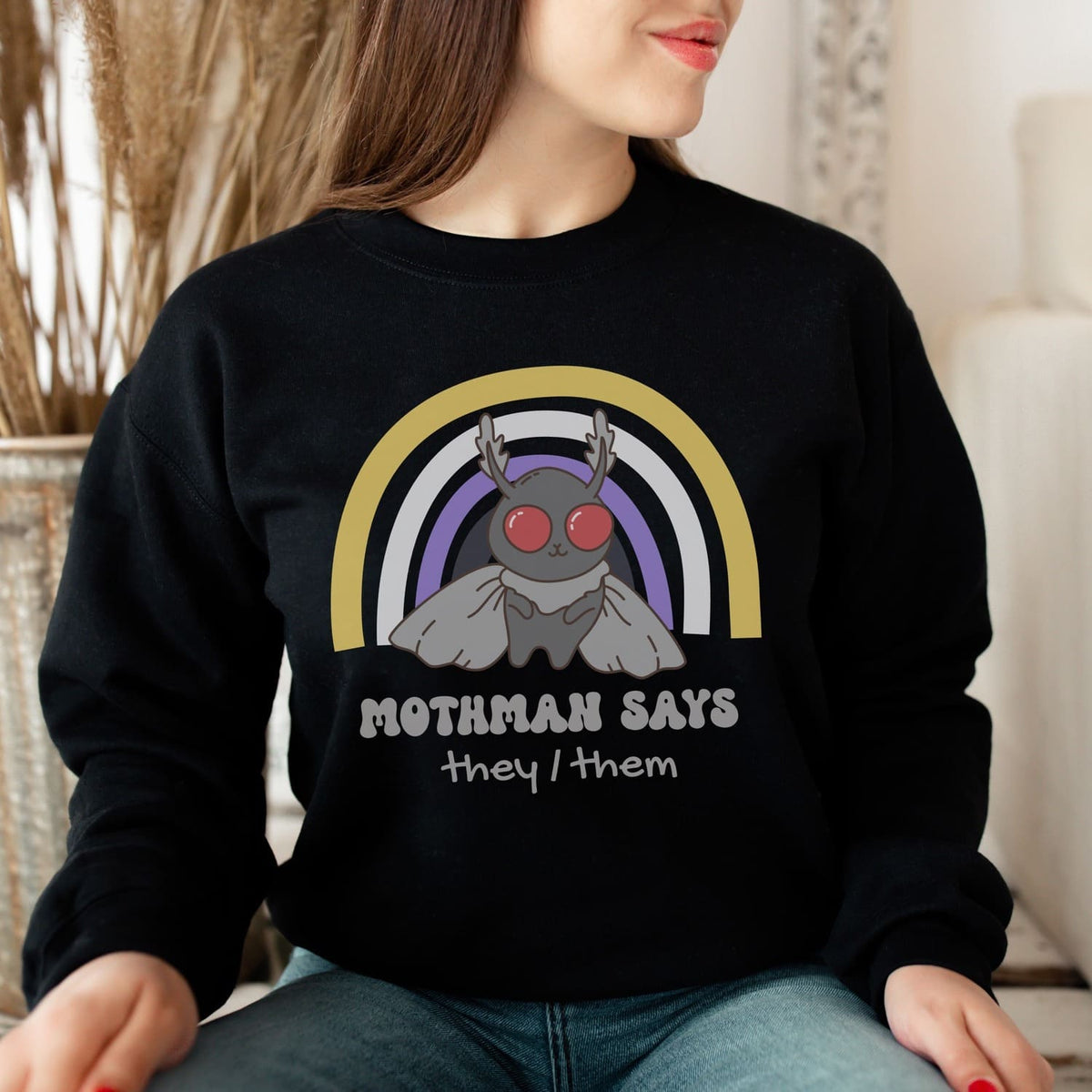 Person wearing a black nonbinary custom pronouns sweatshirt with mothman design and text, sitting indoors.