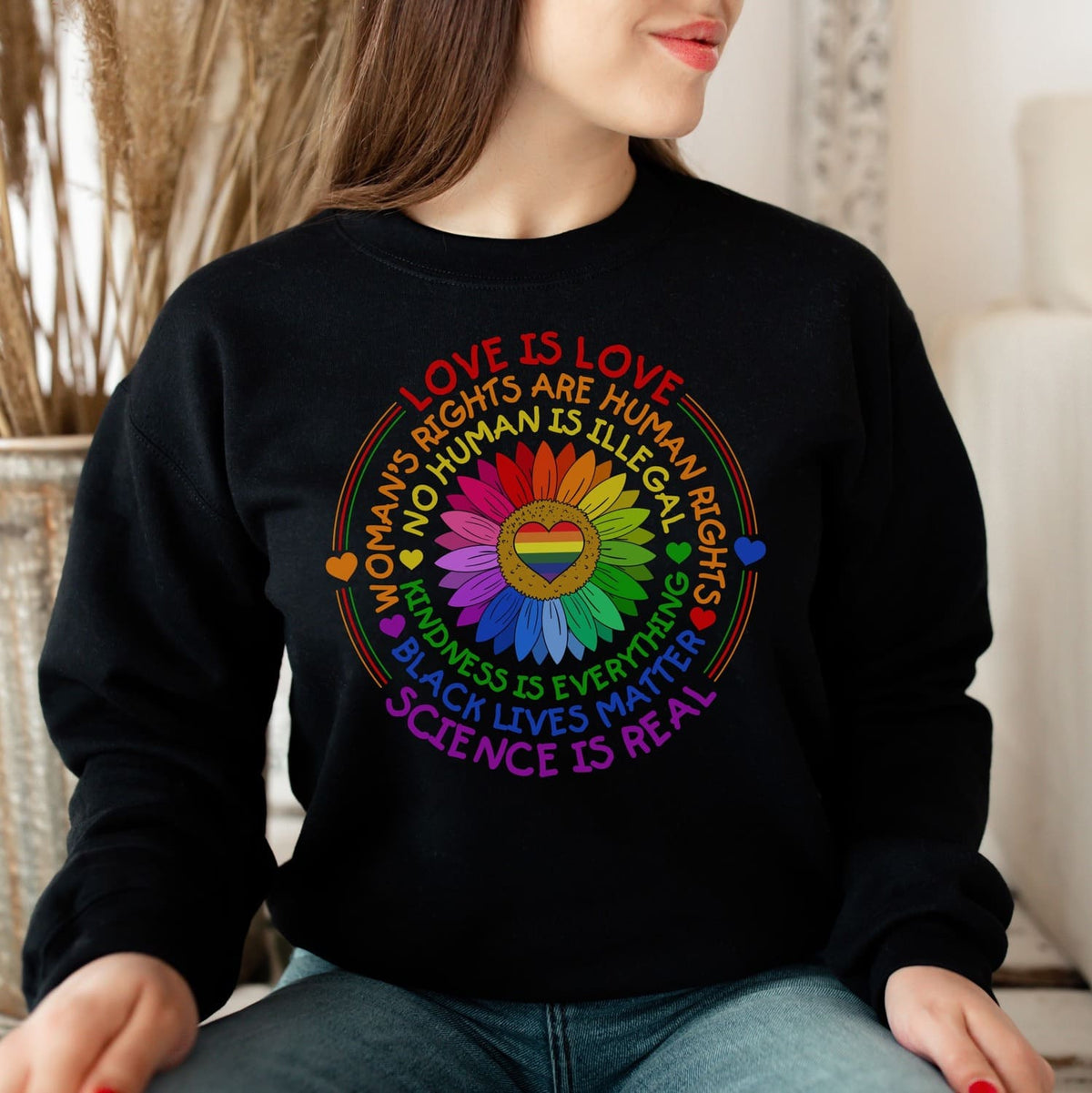 Person wearing a black pride sweatshirt with colorful text and symbols on a blurred indoor background