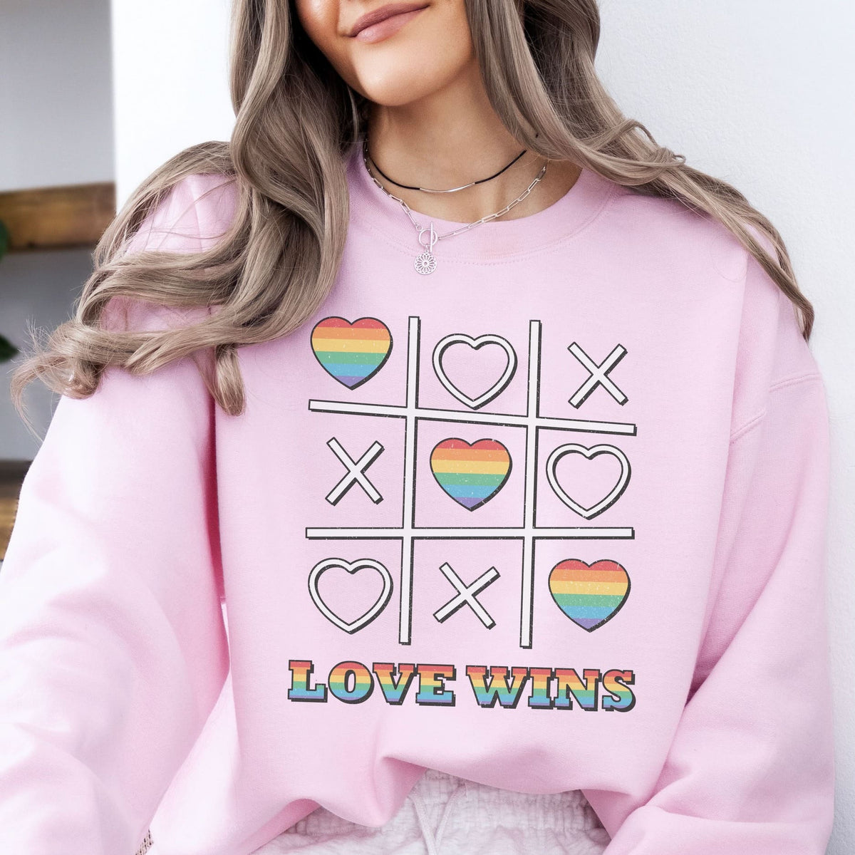 Person wearing a pink LGBTQ pride sweatshirt with a rainbow heart and cross design and 'LOVE WINS' text.