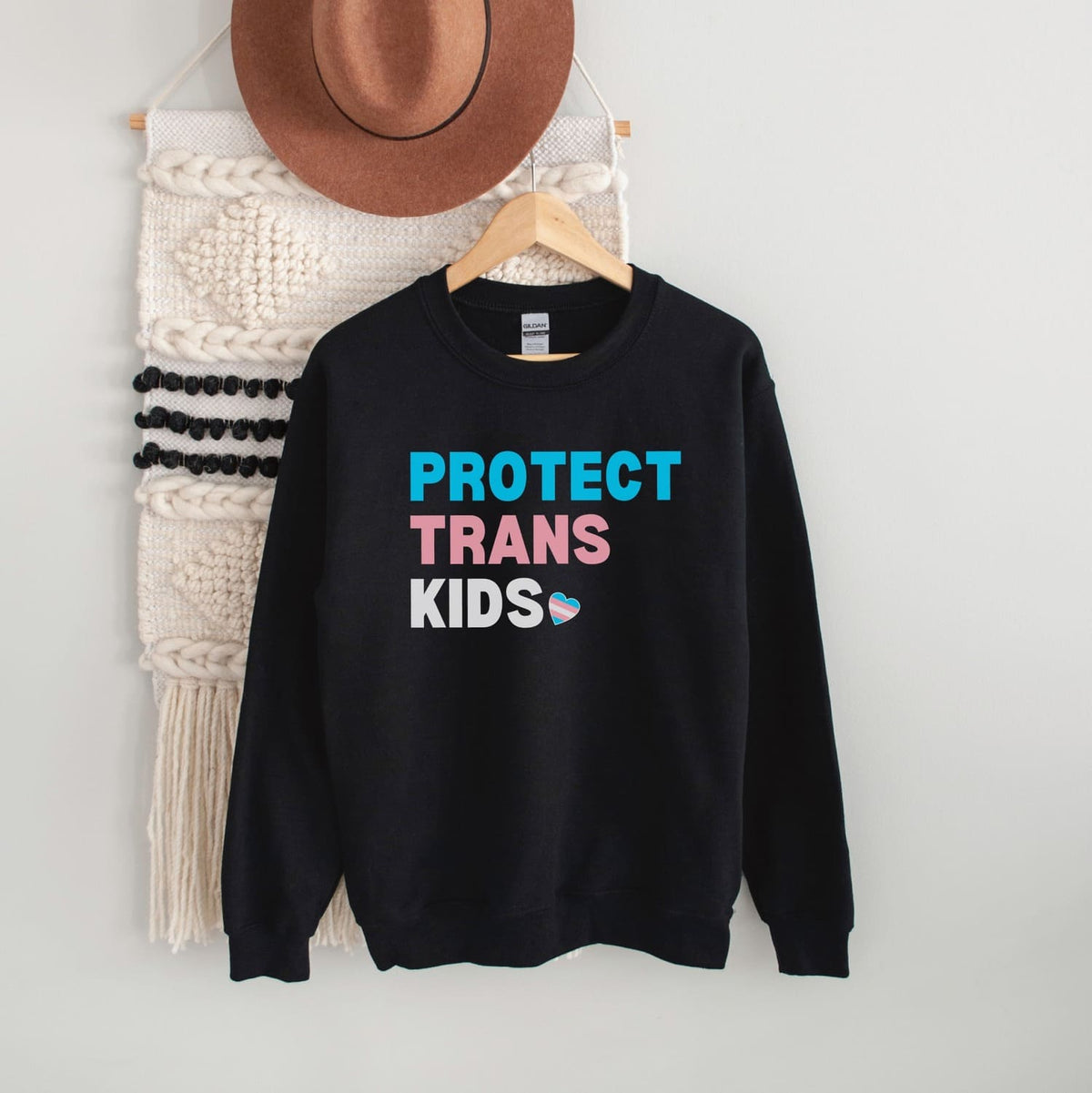 Black transgender sweatshirt with 'PROTECT TRANS KIDS' text on a hanger against a white wall.