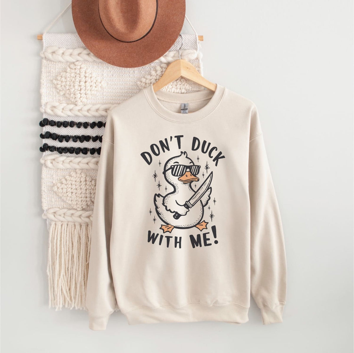 Beige sweatshirt with a funny duck graphic design and text, hanging on a wooden hanger against a white wall.