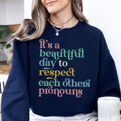 Person wearing a navy pronouns sweatshirt with colorful text, sitting indoors.