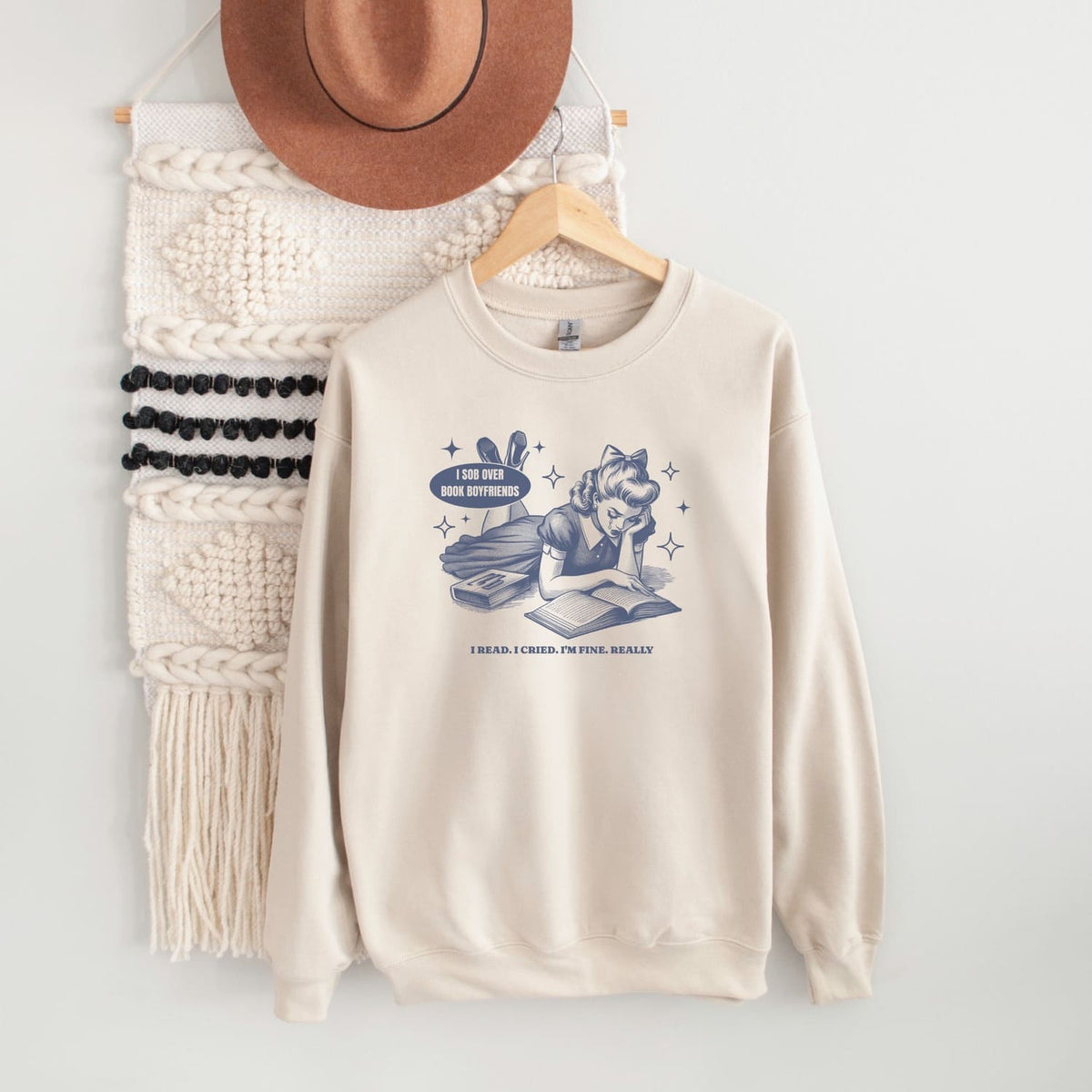 Beige sweatshirt with bookish graphic design on a hanger against a white wall.