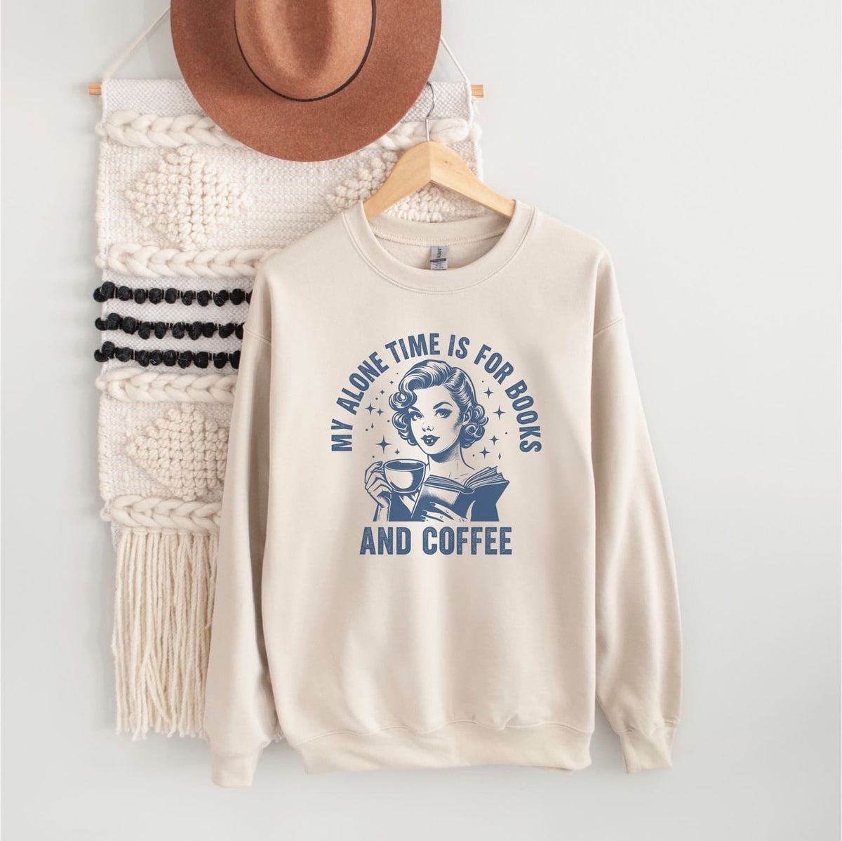 Beige bookish sweatshirt with graphic design and text on a white wall with decor items.