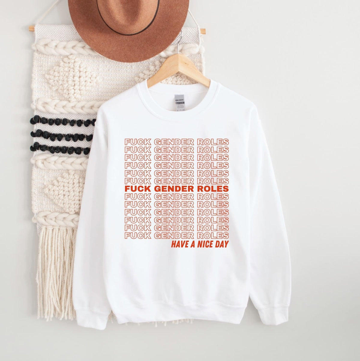 White non cisgender sweatshirt with red text on a hanger next to a knitted item and hat.