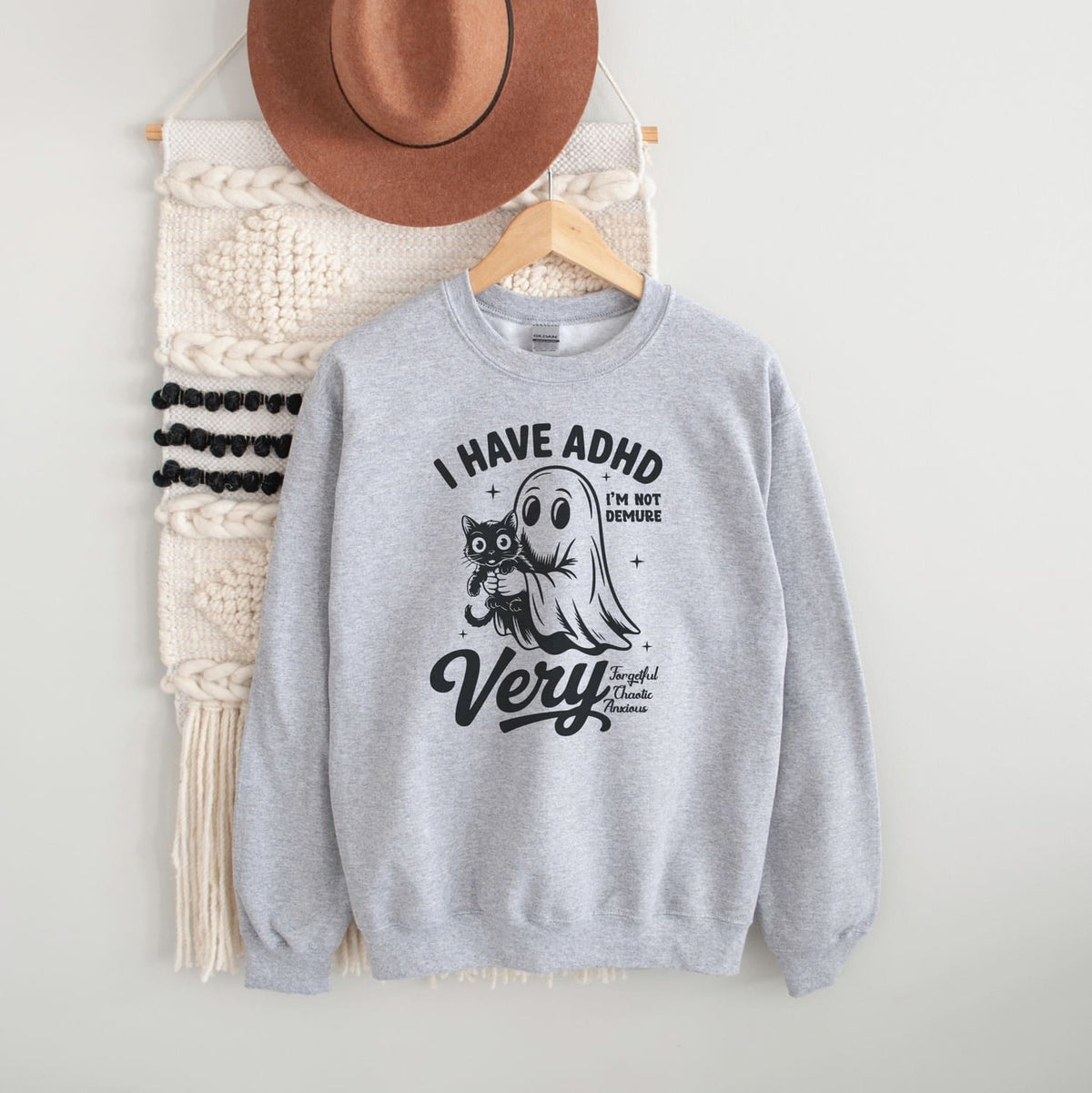 Gray sweatshirt with spooky ADHD design and text, placed on a white surface with a hat and bag in the background.