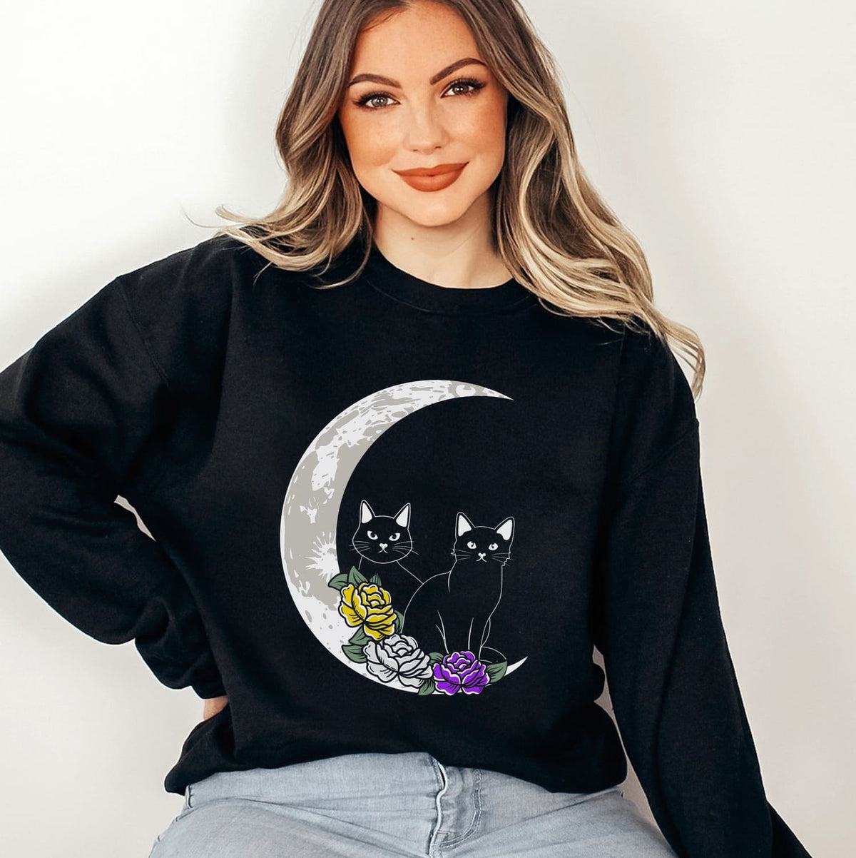 Enby person wearing a nonbinary sweatshirt with a cat and moon design on a plain background
