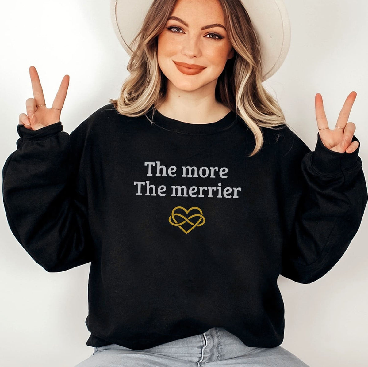 Person wearing a black polyamory sweatshirt with 'The more The merrier' text and heart design, making peace signs.