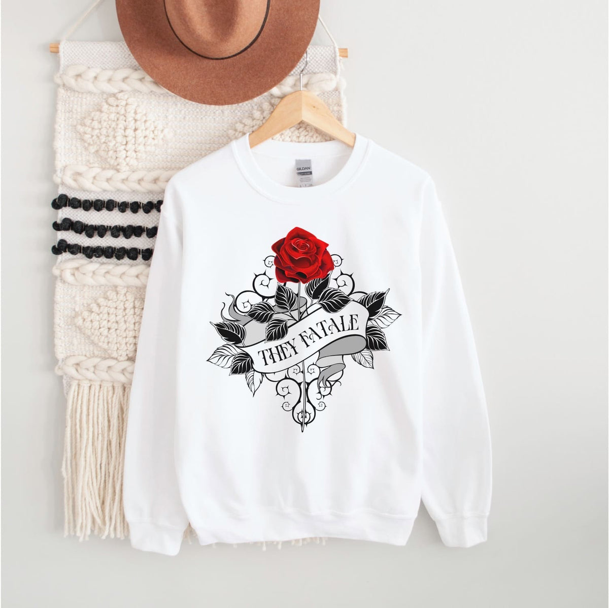 White nonbinary sweatshirt with red rose and 'They Fatale' feminist design on a hanger with a hat and scarf.