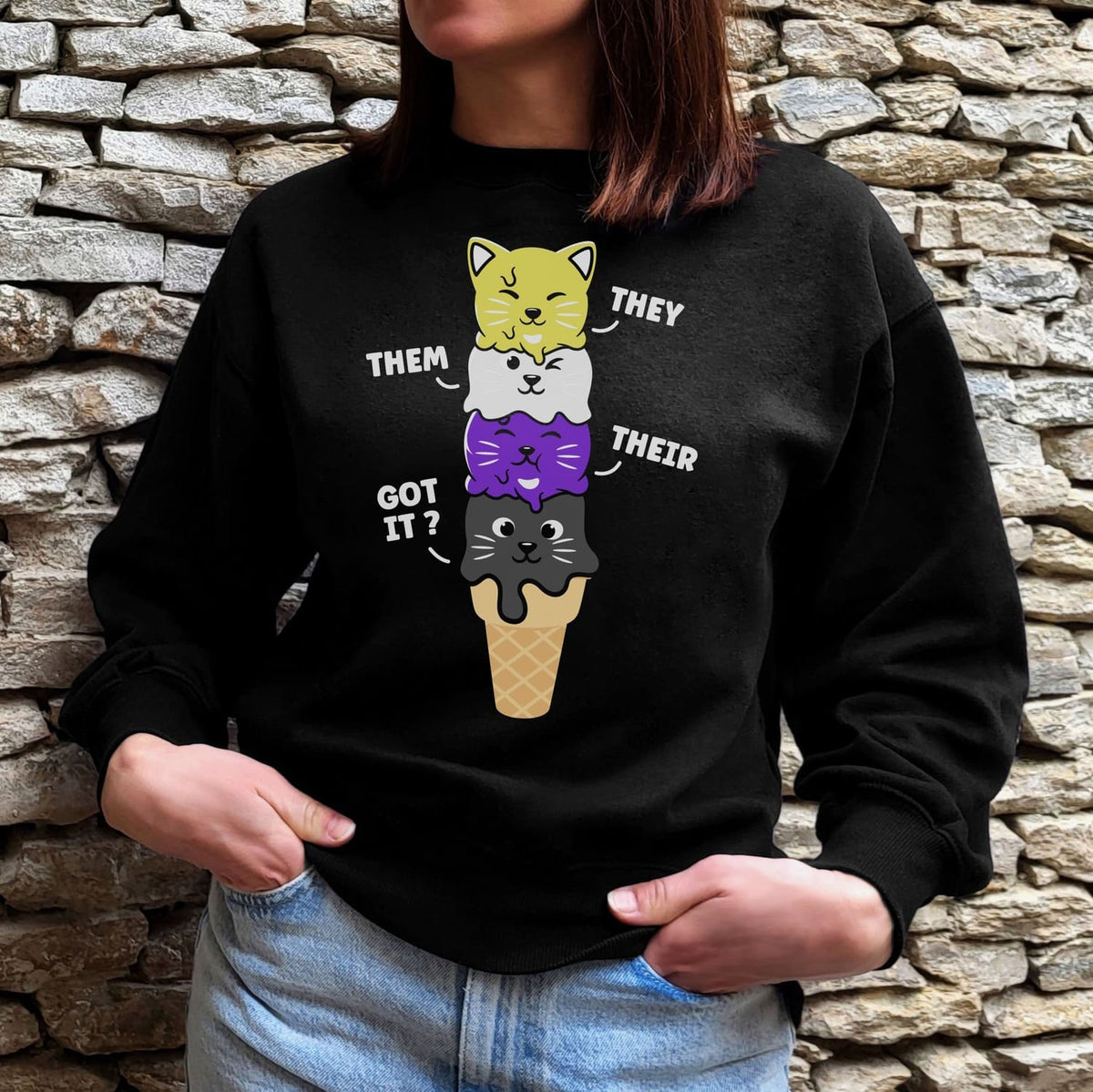 Enby wearing a black sweatshirt with a cute ice cream cone graphic and pronouns, standing against a stone wall.