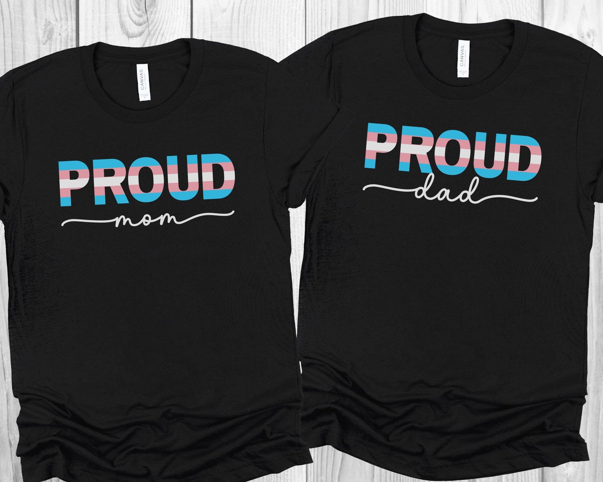 Two black transgender supporting t-shirts with 'PROUD mom' and 'PROUD dad' text on a wooden surface.