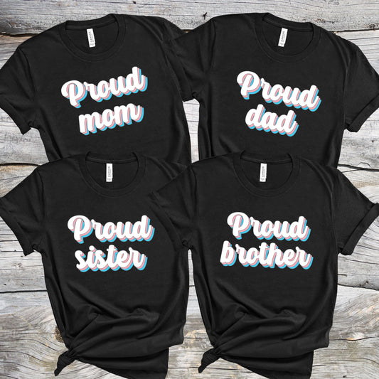 Matching transgender family  t-shirts with 'Proud mom', 'Proud dad', 'Proud sister', and 'Proud brother' text on a wooden surface.