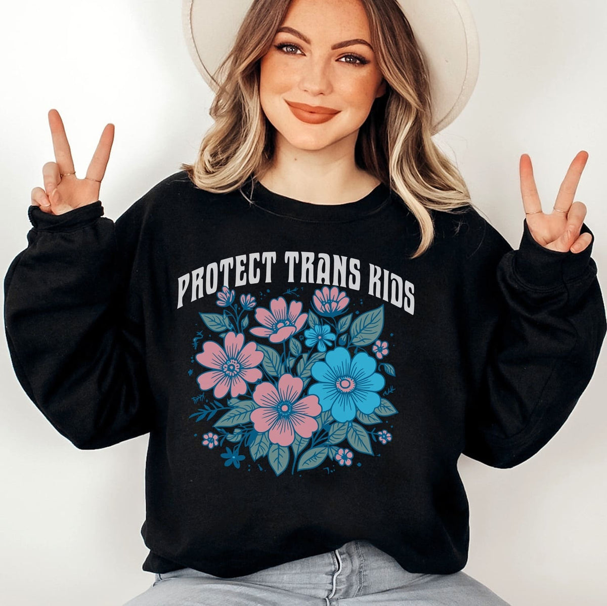 Person wearing a black transgender sweatshirt with floral design and 'PROTECT TRANS KIDS' text, making peace signs.