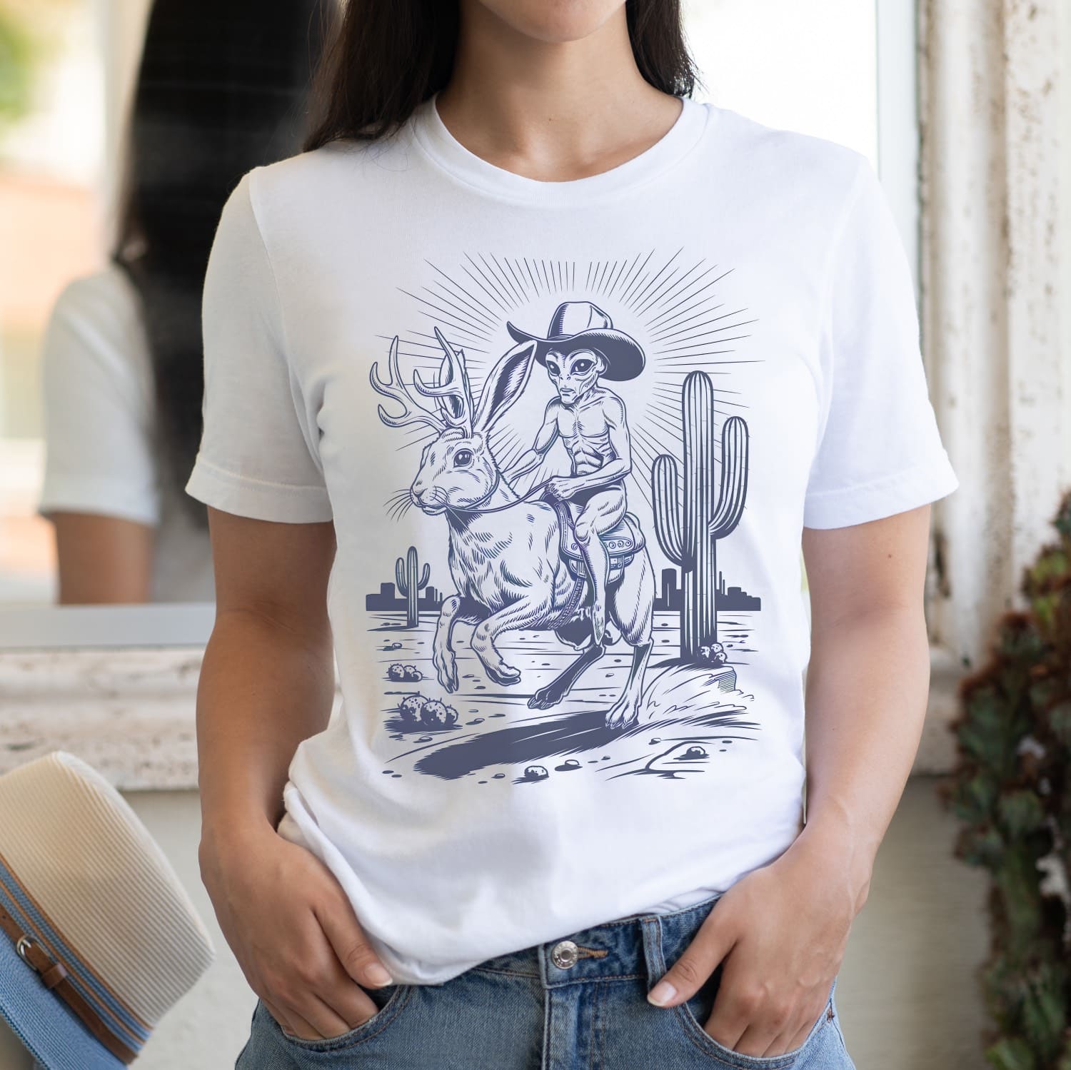 Person wearing a white t-shirt with a graphic design of an alien riding a jackalope in a desert landscape.