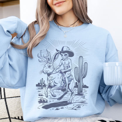 Person wearing a light blue sweatshirt with a graphic design of an alien riding a jackalope next to a cactus.