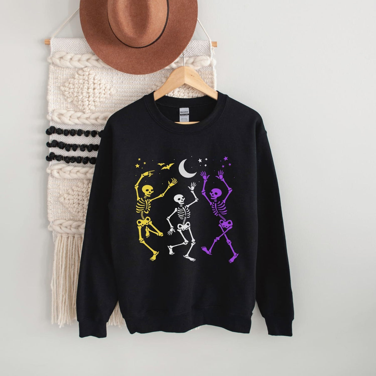 Black sweatshirt with nonbinary dancing skeletons on a hanger against a white wall.